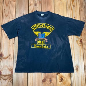 Op de afbeelding: Een marineblauw t-shirt met een geel en rode afbeelding. De afbeelding bevat de woorden "Fifth Chapter" boven een gevleugeld embleem en "M.C. Bass Lake" eronder. Het shirt ligt plat op een houten oppervlak.