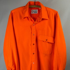 May include: Bright orange long-sleeve button-down shirt with a single chest pocket. The shirt has a classic collar and buttoned cuffs. The brand logo "Sports Afield" is visible on the hanger. A classic, casual garment.