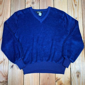 Puede incluir: Un suéter de pana azul real con cuello en V y puños y dobladillo acanalados. El suéter está hecho de una tela suave y texturizada y se exhibe sobre una superficie de madera.