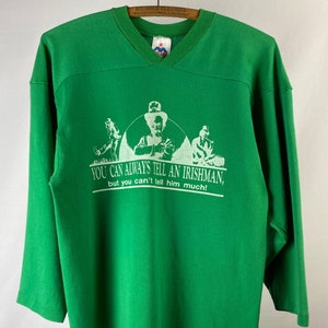 Könnte beinhalten: Grünes Langarmshirt mit V-Ausschnitt und weißer Grafik. Die Grafik zeigt drei Figuren in einem Kreis mit dem Text "YOU CAN ALWAYS TELL AN IRISHMAN, but you can't tell him much!" Ein weißes "L"-Etikett befindet sich unten.