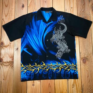 May include: Black short-sleeve button-down shirt with a blue flame design and a white dragon graphic. The shirt has a pointed collar and a series of buttons down the front. A yellow tribal design is at the bottom.