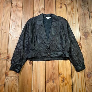 May include: A black cropped jacket featuring a quilted texture on the sleeves and front panels, a notched collar, long sleeves with buttoned cuffs, and a single front button closure. The garment has a structured fit with a defined waistband.