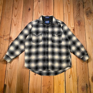 May include: A black and white plaid button-down shirt with two chest pockets. The shirt has long sleeves and a classic collar. The fabric appears to be a wool blend. The shirt is laid flat on a wooden surface.