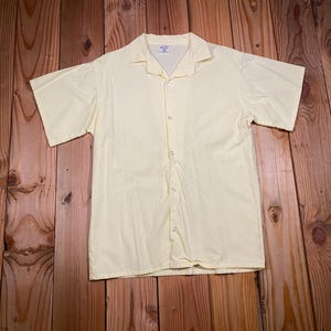 May include: A light yellow short-sleeved button-down shirt with a classic collar and a front button closure. The shirt features a plain, solid-colored design with a standard hemline and a visible brand label located at the inside of the neckline.