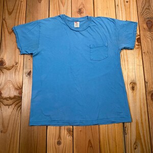 May include: A solid light blue short-sleeve t-shirt featuring a crew neckline and a single patch pocket on the left side of the chest. The garment has a standard fit with a straight hemline and a visible brand label inside the collar area.