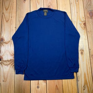 Op de afbeelding: Een effen koningsblauw shirt met lange mouwen, een ronde hals en geribbelde boorden. Het kledingstuk heeft een zichtbaar label in de nek met tekst. Het shirt is gemaakt van een gladde, lichtgewicht stof met een consistente diepblauwe kleur.