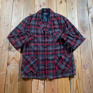 May include: A long-sleeve button-up jacket featuring a red and dark gray plaid pattern. The garment includes a notched collar, a single chest pocket on the left side, and two large patch pockets at the bottom front. The fabric has a textured finish.