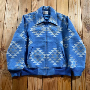 May include: A blue zip-up jacket featuring a repeating geometric diamond pattern in light gray. The jacket has a pointed collar, long sleeves with ribbed cuffs, a ribbed hem, and two front pockets. A white label is visible inside the collar.