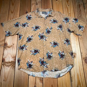 May include: Tan button-down short-sleeve shirt with a repeating abstract pattern in blue, white, and burgundy. The shirt is laid flat on a wooden surface, showcasing its design and style.