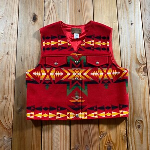May include: A red vest featuring a geometric pattern in yellow, black, green, and white. The garment has a V-neckline, a button-front closure with silver-toned metal buttons, and two chest pockets with buttoned flaps. The interior is lined in red.