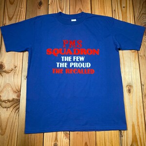 Può includere: Una maglietta blu con la scritta "PMS SQUADRON THE FEW THE PROUD THE RECALLED" in rosso e bianco. La maglietta è appoggiata su una superficie di legno.