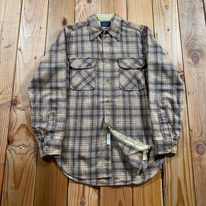 May include: A long-sleeve button-up shirt featuring a brown, tan, and grey plaid pattern. The shirt includes a pointed collar, two chest pockets with button flaps, and a light green interior lining visible at the collar and the partially open hem.