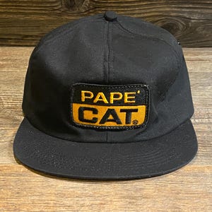 Può includere: Un cappellino da baseball nero con una toppa rettangolare sul davanti. La toppa ha uno sfondo nero con il testo giallo PAPE' sulla riga superiore e il testo giallo CAT sulla riga inferiore, racchiuso in un bordo rettangolare giallo.