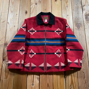 May include: A red zip-up jacket featuring a black collar and a geometric pattern in black, blue, and cream. The design includes horizontal stripes across the chest and diamond and triangular motifs along the sleeves and the lower hem area.
