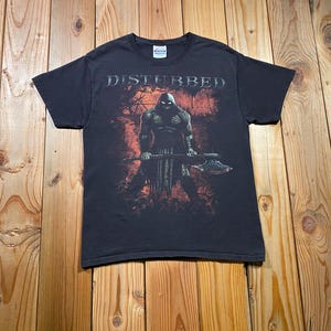 May include: A black short-sleeved t-shirt featuring the word DISTURBED in white lettering at the top. The central graphic depicts a hooded, muscular figure in dark armour holding a large battle axe against a distressed red and black background.