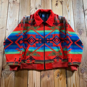 May include: A long-sleeve zip-up jacket featuring a vibrant geometric pattern in shades of red, blue, green, and black. The jacket has a solid red collar, ribbed cuffs, a ribbed hem, and two front pockets with a black zipper closure down the center.