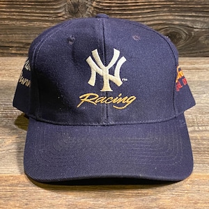 May include: A navy blue baseball cap featuring an embroidered white New York Yankees logo on the front with the word Racing in gold script underneath. The cap includes additional embroidered text details on the side panels in gold and white thread.