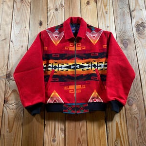 May include: A red wool jacket featuring a vibrant geometric pattern in orange, yellow, black, and white. The jacket has a full-length front zipper, a fold-down collar, and black ribbed knit cuffs and hem. The design includes repeating angular shapes.