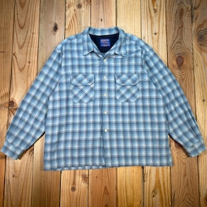 Puede incluir: Una camisa de manga larga con botones y un patrón de cuadros en azul claro y blanco. La camisa incluye un cuello puntiagudo, un forro interior azul oscuro y dos bolsillos en el pecho con solapas abotonadas. Se cierra con una fila de botones.