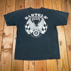 May include: Dark gray t-shirt with a graphic on the back that reads "Bartels Custom Bike Show" with an eagle, checkered flags, and a wheel. The text "Marina del Rey, CA" is also included.