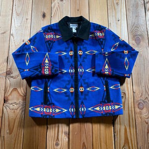 May include: A blue zip-up jacket featuring a geometric pattern in red, yellow, and black. The jacket has a solid black collar, a full-length front zipper, and two vertical side pockets. A Pendleton brand label is visible on the inner neck area.