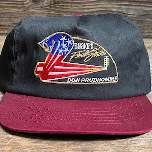May include: Black and burgundy baseball cap with a flat brim. The cap features an embroidered patch with the text "Snake's Final Strike" and a design incorporating the American flag. The text "DON PRUDHOMME" is also visible.
