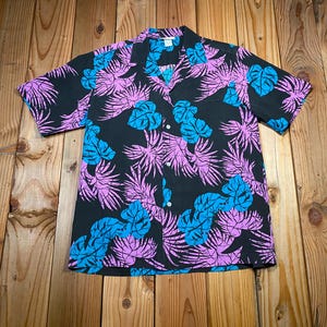 Puede incluir: Una camisa de manga corta con botones en color negro que presenta un vibrante estampado de hojas tropicales en tonos rosa brillante y azul cian. La prenda tiene un cuello clásico, un dobladillo recto y botones blancos en la parte central.