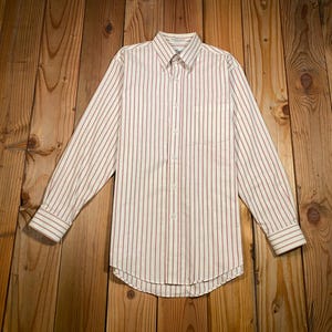 May include: A long-sleeve button-down shirt featuring a vertical striped pattern in red and dark blue on a white background. The shirt includes a pointed collar, a single chest pocket on the left side, and buttoned cuffs on the long sleeves.