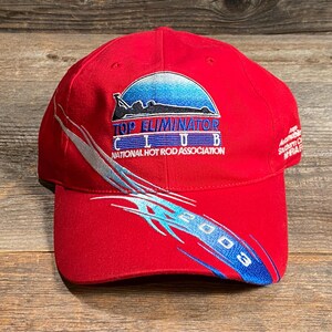 Op de afbeelding: Een felrode baseballpet met het geborduurde logo van de Top Eliminator Club National Hot Rod Association. Het ontwerp bevat een blauwe gradi&euml;nt halve cirkel, een zwart silhouet van een raceauto en blauwe tribale graphics met 2003.