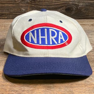 Op de afbeelding: Een witte baseballcap met een marineblauwe klep. De cap toont de letters "NHRA" in blauw, binnen een rode ovaal, op een witte achtergrond. De cap heeft een marineblauwe knop bovenop.