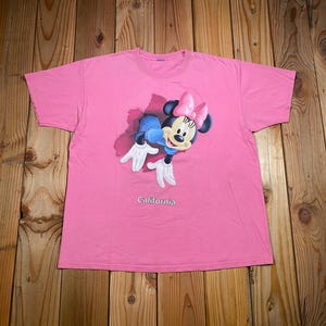 May include: A bright pink short-sleeved t-shirt featuring a graphic of Minnie Mouse emerging from a dark red cutout shape. Minnie Mouse wears a pink bow and white gloves. The word California is printed in white text below the graphic.