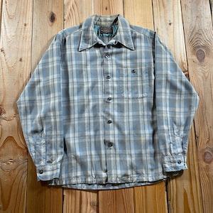 Puede incluir: Una camisa de manga larga con botones y un patrón de cuadros en azul claro, gris y blanco. La camisa incluye un cuello puntiagudo, un bolsillo en el pecho en el lado izquierdo y botones a lo largo de la parte frontal y en los puños.