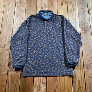 May include: A long-sleeve polo shirt in a dark blue hue featuring an all-over gold and tan paisley pattern. The garment includes a solid dark blue collar, a three-button placket, and ribbed cuffs at the end of each long sleeve.