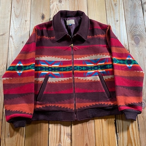 Peut inclure: Une veste zippée présentant un motif à rayures horizontales dans des tons bordeaux, rouge et orange. Le vêtement comprend un motif géométrique sur la poitrine en bleu, vert et blanc, un col foncé uni, ainsi que des poignets et un ourlet côtelés.