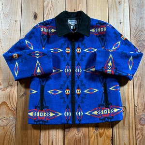 May include: A blue zip-up jacket featuring a geometric pattern in red, yellow, black, and white. The jacket has a solid black collar, a full-length black front zipper, and two vertical zippered pockets. A Pendleton brand label is visible at the neck.