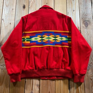 May include: A bright red jacket featuring a horizontal band across the back with a geometric pattern in blue, green, yellow, and dark red. The jacket has long sleeves, a ribbed knit hem, and ribbed knit cuffs, with a collar at the neckline.