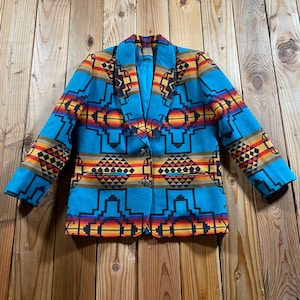 Puede incluir: Un blazer de color turquesa brillante con un llamativo patrón geométrico en tonos naranja, amarillo, rojo y negro. La chaqueta tiene un cuello chal ancho, mangas largas, dos bolsillos delanteros y un cierre de botones en el centro.