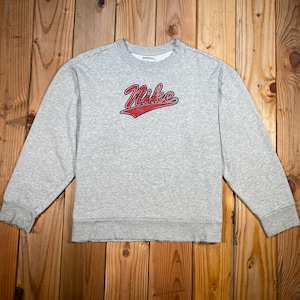 Puede incluir: Una sudadera gris jaspeada con el logo de Nike en rojo. La parte superior de manga larga y cuello redondo est&aacute; hecha de un material suave. La sudadera se muestra sobre una superficie de madera.