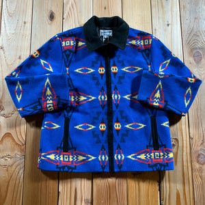 May include: A blue zip-up jacket featuring a geometric pattern in red, yellow, and black. The jacket has a solid black collar and black trim along the vertical zipper and two side pockets. A Pendleton brand label is visible at the inner neckline.