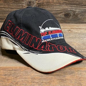 May include: Black baseball cap with "ELIMINATOR" in red lettering. The cap features a white bill with red trim and the text "TOP ELIMINATOR CLUB NATIONAL HOT ROD ASSOCIATION". The hat is likely a collectible item.
