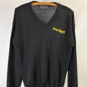 Puede incluir: Suéter negro con cuello en V y mangas largas. El suéter tiene dobladillo y puños acanalados. Las palabras "Shop Smart Food Warehouse" están impresas en amarillo en la parte superior izquierda del pecho. El suéter está colgado en una percha de madera.