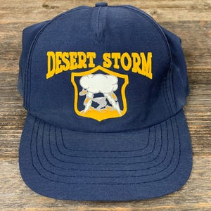 Pode incluir: Boné de beisebol azul marinho com as palavras "DESERT STORM" em letras amarelas. O boné apresenta um emblema amarelo e branco de uma nuvem com raios. O boné é feito de um tecido durável.