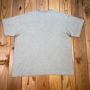 Puede incluir: Una camiseta gris jaspeada de manga corta, colocada sobre una superficie de madera. La camiseta tiene cuello redondo y un dise&ntilde;o sencillo y cl&aacute;sico. La tela parece ser una mezcla de algod&oacute;n suave.