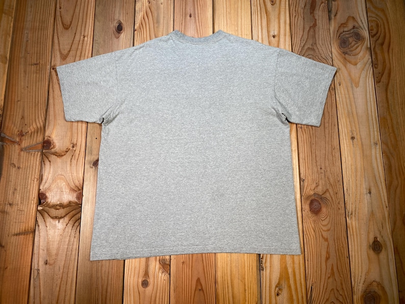 Puede incluir: Una camiseta gris jaspeada de manga corta, colocada sobre una superficie de madera. La camiseta tiene cuello redondo y un dise&ntilde;o sencillo y cl&aacute;sico. La tela parece ser una mezcla de algod&oacute;n suave.