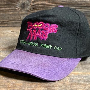 May include: Black baseball cap with a purple brim. The cap features the text "Boogie Man" in pink and "Federal-Mogul Funny Car" in green.
