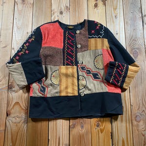 May include: A long-sleeved jacket featuring a patchwork design in black, red, tan, and brown. The garment includes embroidered floral and geometric patterns, a round neckline, and a front button closure with five circular buttons down the centre.