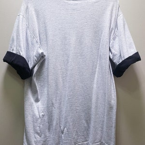 May include: A gray short-sleeved t-shirt with black trim on the sleeves. The shirt is hanging on a hanger.