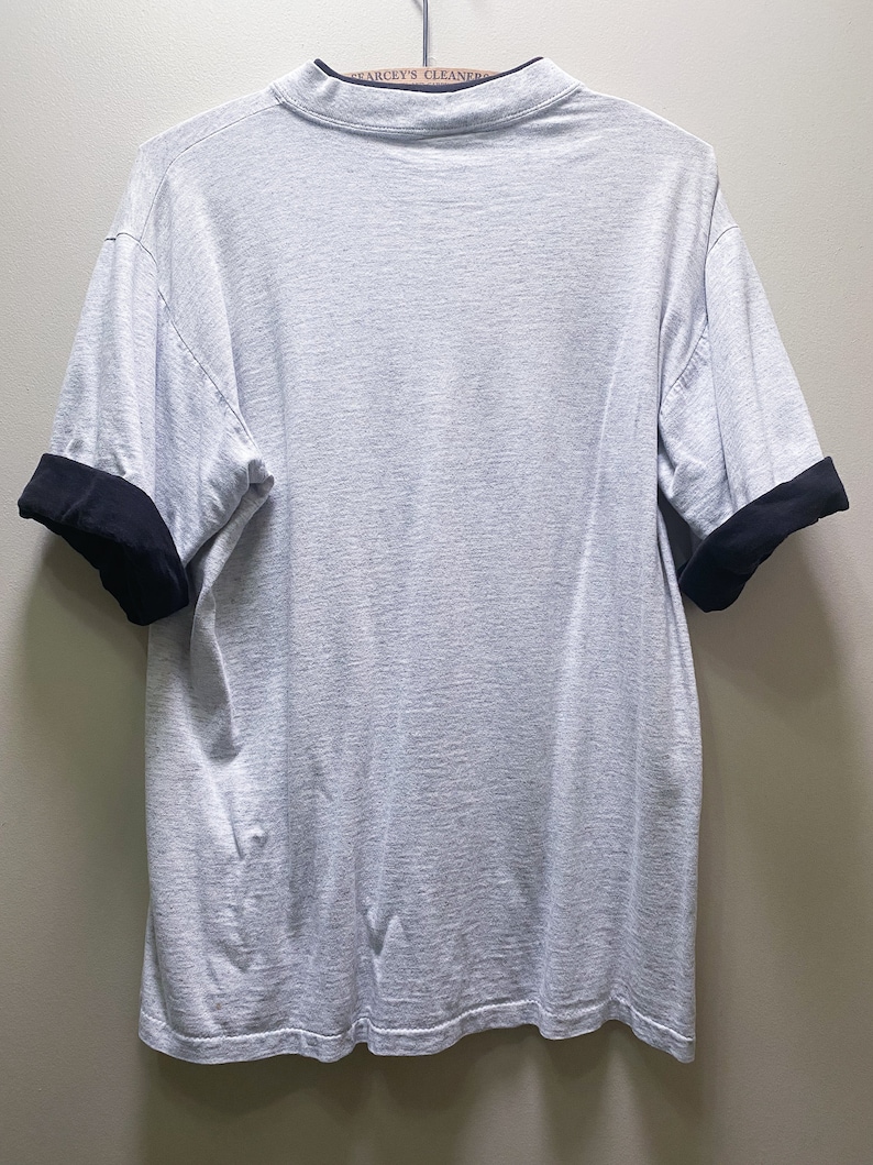 May include: A gray short-sleeved t-shirt with black trim on the sleeves. The shirt is hanging on a hanger.