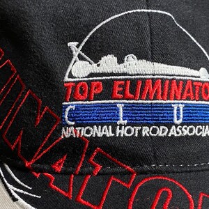 May include: Black baseball cap with red and white embroidered text and graphics. The cap features the words "ELIMINATOR" and "TOP ELIMINATOR CLUB" with a graphic of a race car. The text "NATIONAL HOT ROD ASSOCIATION" is also visible.