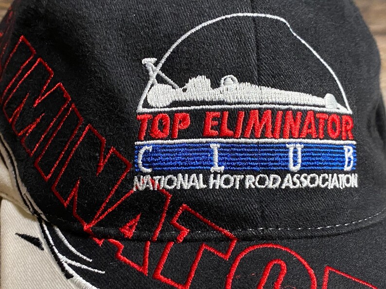 May include: Black baseball cap with red and white embroidered text and graphics. The cap features the words "ELIMINATOR" and "TOP ELIMINATOR CLUB" with a graphic of a race car. The text "NATIONAL HOT ROD ASSOCIATION" is also visible.